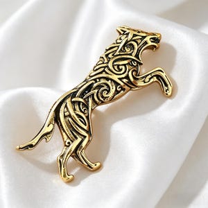 May include: A gold-colored metal brooch in the shape of a wolf in a rearing pose. The wolf has intricate Celtic knotwork detailing throughout its body. The brooch is set against a white, silky background.