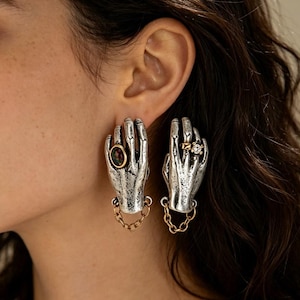 Può includere: Un paio di orecchini color argento a forma di mano, ciascuno con una piccola catenella color oro al polso. Una mano indossa un anello con una pietra ovale scura, mentre l'altra mano indossa un anello color oro con una pietra rotonda trasparente.