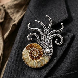 May include: A brooch featuring a polished, spiral-patterned brown and tan ammonite fossil shell base attached to a textured silver-toned metal octopus head with multiple curved, detailed tentacles extending upward from the fossilized shell.