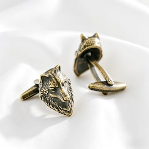 May include: A pair of antique brass cufflinks featuring detailed wolf head designs. The cufflinks have a textured finish and a classic T-bar closure. The wolf heads are facing different directions, adding visual interest to the accessory.
