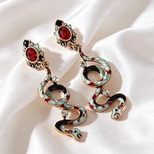 May include: Gold-coloured snake earrings. Each earring has a red gemstone and a serpentine design with light blue, red, and black enamel. Clear crystals are set along the snake's body. The earrings are on a white background.