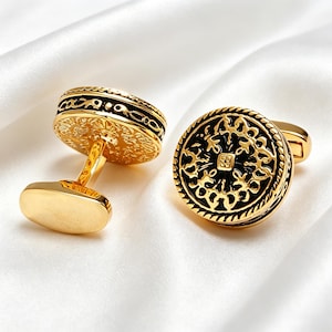 Könnte beinhalten: Goldfarbene Manschettenknöpfe mit aufwendigen schwarzen und goldenen Filigranmustern. Die Manschettenknöpfe haben eine runde Oberfläche mit einem detaillierten Muster und einen rechteckigen Steg. Das Design ist vor einem weißen Hintergrund.