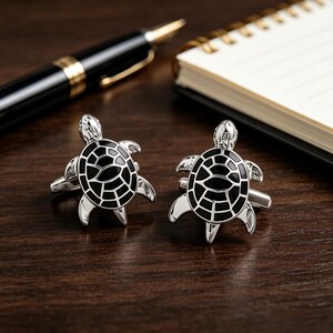 May include: A pair of silver cufflinks with a black enamel turtle design. The cufflinks are on a dark wooden surface, with a pen and notepad in the background. The turtle design features a detailed shell pattern.