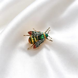 Könnte beinhalten: Goldfarbene Insektenbrosche mit grünen, roten und schwarzen Emaille-Details. Die Brosche zeigt ein detailliertes Design eines Käfers mit komplizierten Flügelmustern und Antennen. Die Brosche ist vor einem weißen Satinhintergrund platziert.