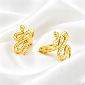 Puede incluir: Dos anillos dorados con forma de serpiente y detalles blancos. Los anillos se muestran sobre una superficie blanca y sedosa. Las serpientes están enrolladas, con la cabeza levantada. Los anillos son probablemente de metal.