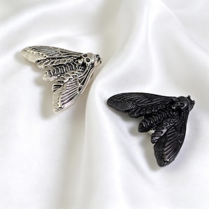 May include: Two insect-shaped brooches. One is silver-toned, the other is black. Both have detailed wing and body designs. The brooches are displayed on a white, silky fabric.