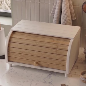 Peut inclure: Une boîte à pain rectangulaire dotée d'un cadre blanc et d'une porte coulissante à rideau composée de lattes en bambou brun clair. L'unité présente un petit bouton rond sur le devant et repose sur quatre pieds courts pour le plan de travail.