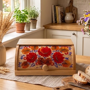 May include: A wooden bread bin with a roll-top door featuring a hand-painted floral design in shades of red, orange, and blue. The bin has a light wood finish and a round wooden knob on the front panel, sitting on a linen runner on a table.