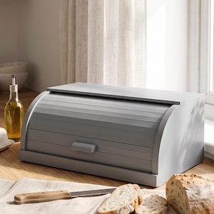 Puede incluir: Una panera rectangular de madera gris con tapa enrollable y un pequeño tirador. La panera presenta listones horizontales en el panel frontal curvo y se asienta sobre una base plana, ofreciendo una solución para guardar el pan.