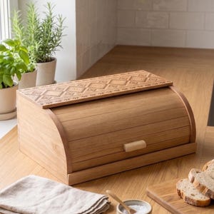 Puede incluir: Una panera de madera de color marrón claro con tapa enrollable que presenta un patrón geométrico de diamantes grabado con láser. El recipiente rectangular tiene un pequeño tirador de madera en el panel frontal y está hecho de madera natural.