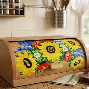 May include: A rectangular wooden bread bin with a roll-top lid featuring a hand-painted floral design. The artwork displays vibrant yellow sunflowers, small white daisies, blue blossoms, and red accents against the natural light brown wood grain.