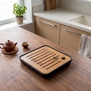 Puede incluir: Una tetera y una taza de cerámica marrón están sobre una mesa de madera, junto a una bandeja de té de bambú. El juego de té está colocado cerca de una ventana y una encimera.