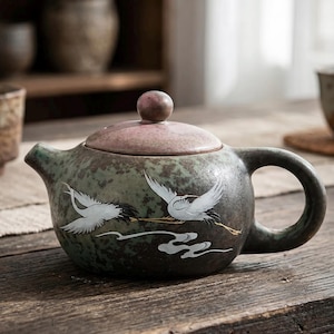 Può includere: Una teiera in ceramica con una glassa verde e marrone screziata. Il coperchio è rosa tenue con un piccolo pomello rotondo. Due gru bianche sono dipinte sul lato, con un design a nuvola stilizzato. La teiera ha un manico e un beccuccio arrotondati.