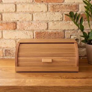 May include: A rectangular wooden bread bin with a curved, roll-top lid and a small horizontal handle centred on the front panel. The container is crafted from light-toned wood with a visible grain pattern and features a smooth, finished surface.