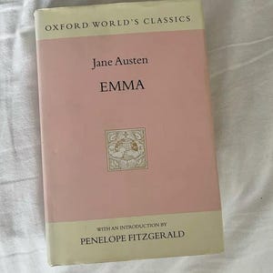 May include: A hardcover book titled "Emma" by Jane Austen, part of the Oxford World's Classics series. The book has a light pink cover with white text and a small illustration. The introduction is by Penelope Fitzgerald.