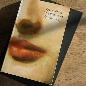 May include: A hardcover book titled "The Picture of Dorian Gray" by Oscar Wilde. The book cover features a close-up of a face with red lips. The book rests on top of a black book on a wooden surface.