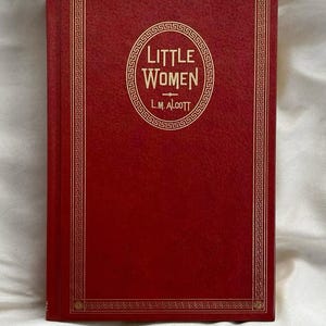 May include: A hardcover book titled "Little Women" by L.M. Alcott. The book has a red cover with gold decorative borders and lettering. The title is in a circular design in the center of the book.