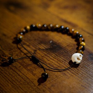 May include: A beaded bracelet featuring round, brown and black tiger eye beads and a single white skull-shaped bead. The bracelet is constructed with a black cord and includes an adjustable sliding knot closure with two small beads at the ends.