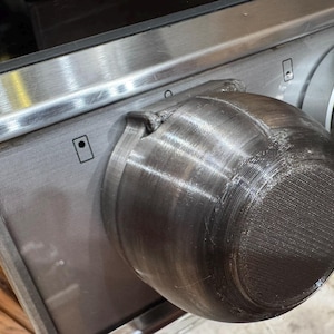 May include: A dark gray, 3D-printed replacement stove control knob with a textured, layered finish. The knob is mounted on a stainless steel control panel featuring black printed icons and a secondary metallic knob labeled with HI and numbers.