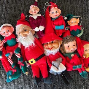 May include: A collection of vintage Christmas ornaments featuring several elf dolls in red and green felt outfits, a Santa Claus figure with a white beard, and a small doll with brown hair, all with painted facial features and attached hanging strings.