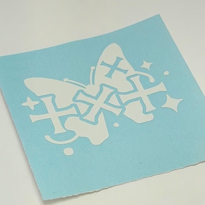 May include: A light blue rectangular decal featuring a white butterfly silhouette. The butterfly design is overlaid with three white cross shapes and accented with small white star and dot motifs, creating a layered graphic appearance.