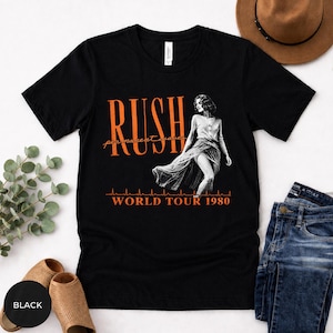 Può includere: T-shirt nera con il nome della band "RUSH" in arancione, le parole "permanent waves" in corsivo e un'immagine in bianco e nero di una donna. Il testo "WORLD TOUR 1980" è anch'esso arancione.