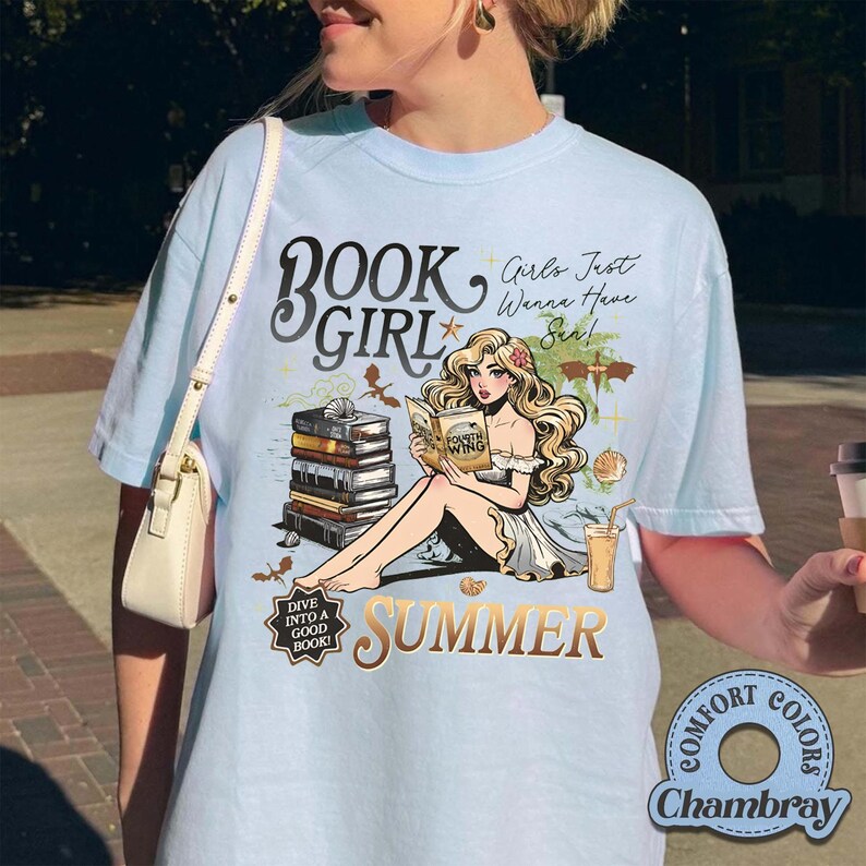 May include: Pale blue t-shirt featuring a graphic of a woman reading, with the words "BOOK GIRL SUMMER" and "Girls Just Wanna Have Sun!". The design includes books, seashells, and a drink. The shirt is labeled "Comfort Colors Chambray".