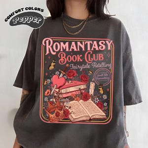 May include: Dark grey t-shirt with a graphic design featuring the text "Romantasy Book Club Fairytale Retelling." The design includes a heart pierced by a sword, books, roses, a crown, and butterflies. The shirt also has the words "Comfort Colors Pepper" printed on it.