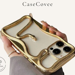 Può includere: Una custodia trasparente per smartphone con un design a rilievo a forma di serpente color oro metallizzato che avvolge i bordi e il retro. La custodia include una cornice dorata rialzata attorno al modulo fotocamera e il testo CaseCovee in alto.