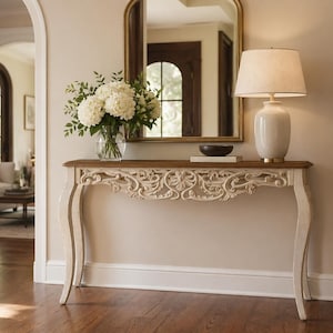 May include: A decorative console table with a carved, off-white apron and curved legs. The table has a dark wood top, holding a lamp, a bowl, and a vase of white flowers. A gold-framed mirror hangs above the table.