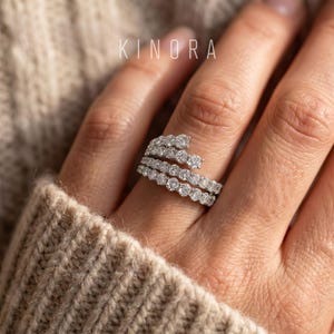 May include: A silver-toned bypass ring featuring a spiral design set with multiple round-cut clear stones. The ring wraps around the finger in a multi-row arrangement. The text KINORA is visible in the upper portion of the image.