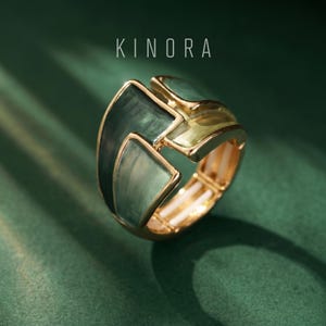 May include: A gold-toned ring with a geometric design featuring shades of green enamel. The ring has a bold, angular shape with the word "KINORA" in white text at the top. The ring is set against a dark green background.