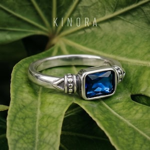 May include: A silver ring with a square, deep blue gemstone. The ring has decorative details on the band and around the stone. The word "KINORA" is printed in white at the top. The ring is displayed on a green leaf.