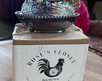May include: An iridescent purple and blue glass hen on nest figurine resting on a cardboard box. The box features the text Rose's Closet, Hen On Nest Exclusive Collection, Timeless Sophistication With Modern Style, and a black rooster graphic.