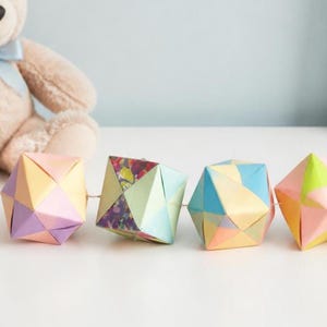 May include: A garland of four geometric paper ornaments strung together. Each ornament is a multi-faceted polyhedron made from folded paper in various pastel shades including pink, yellow, blue, purple, and green, with one featuring a floral pattern.