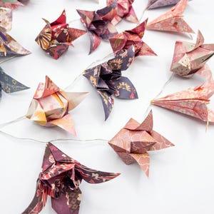 May include: A garland of origami flower lights in shades of pink, purple, and burgundy. The paper flowers are strung on a thin white wire, creating a decorative lighting feature. The flowers display detailed patterns.
