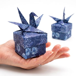 May include: Two origami gift boxes, one held in a hand, are featured. The boxes are navy blue with a white floral pattern. Each box is topped with a folded paper crane. The background is white.