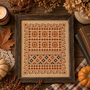 Könnte beinhalten: Ein gerahmtes Kreuzstich-Muster auf beigem Stoff mit einem sich wiederholenden Muster aus kleinen orangefarbenen Kürbissen und dunkelgrünen Blumenmotiven. Das Design ist von einer dekorativen Bordüre aus orangefarbenen Blättern und Eicheln umgeben und in einem dunklen Holzrahmen gefasst.