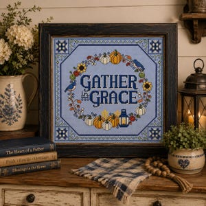 May include: A framed cross-stitch sampler featuring the text GATHER in GRACE in dark blue, surrounded by a wreath of pumpkins, sunflowers, and blue birds on a light blue background, all enclosed in a dark, distressed wooden frame.