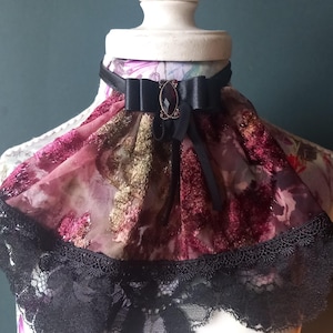 May include: A decorative choker necklace with a black satin ribbon and bow. The choker features a floral patterned fabric in shades of pink, burgundy, and gold, with a black lace trim. A dark jewel is centered on the bow.