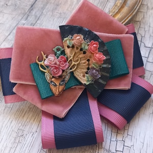 May include: A pink velvet bow with a navy blue ribbon and a decorative floral brooch featuring a deer, flowers, and a fan.