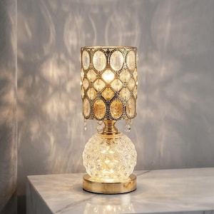 May include: A table lamp featuring a cylindrical shade with a gold-toned metal lattice frame and embedded oval clear crystals. The base is a spherical, textured clear glass orb resting on a circular gold-toned metal stand, emitting a warm light.