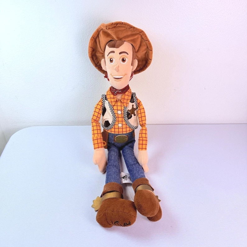 Puede incluir: Un mu&ntilde;eco de peluche de un personaje vaquero que lleva un sombrero marr&oacute;n, una camisa de manga larga a cuadros amarillos y naranjas, un chaleco blanco con manchas negras, pantalones de mezclilla azules y botas marrones con espuelas doradas.