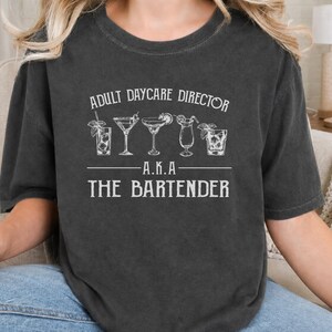 Pode incluir: Uma t-shirt de manga curta cinzento-carvão com texto serigrafado em branco que diz ADULT DAYCARE DIRECTOR A.K.A THE BARTENDER, acompanhada por uma linha de cinco ilustrações distintas de copos de cocktail, incluindo um martini e uma margarita.
