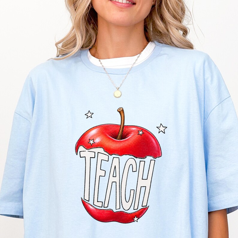 May include: Light blue t-shirt with a large red apple graphic. The apple is cut open to reveal the word "TEACH" in white block letters. Four white stars surround the apple. The shirt has a white crew neck collar.