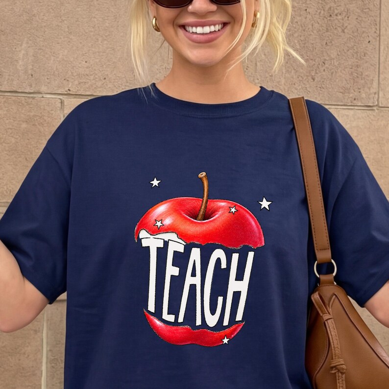 May include: Navy blue t-shirt featuring a red apple graphic with the word "TEACH" in white block letters. The apple design includes a bite taken out of it, with three white stars around the apple. A brown shoulder bag is visible.