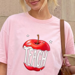 May include: A light pink t-shirt featuring a graphic of a red apple with the word "TEACH" in white block letters. The apple design includes a brown stem and is surrounded by small white stars. The shirt is a crew neck style.