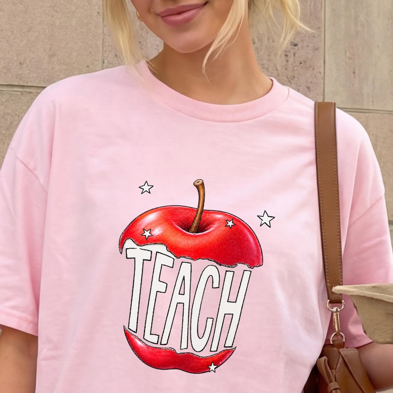 May include: A light pink t-shirt featuring a graphic of a red apple with the word "TEACH" in white block letters. The apple design includes a brown stem and is surrounded by small white stars. The shirt is a crew neck style.