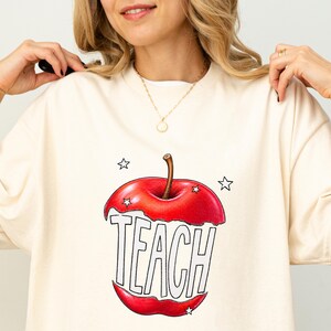 May include: Cream-colored sweatshirt featuring a large red apple graphic with the word "TEACH" in bold, outlined letters. The apple design includes star accents. A gold necklace is visible.