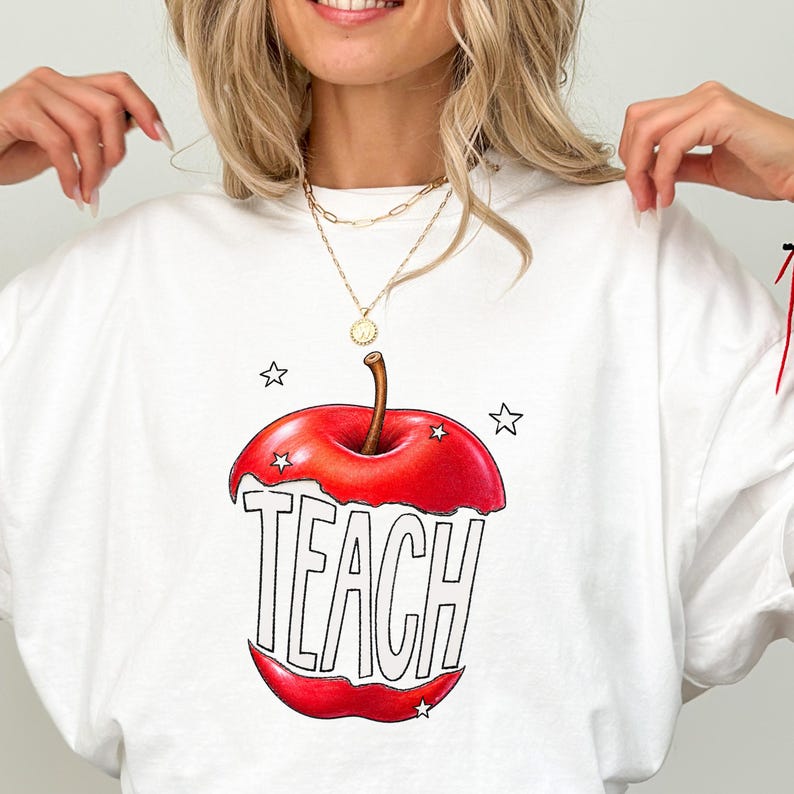 May include: White crew neck t-shirt featuring a large red apple graphic with the word "TEACH" in bold letters. The apple design includes a brown stem and is surrounded by small white stars. A gold necklace is visible.